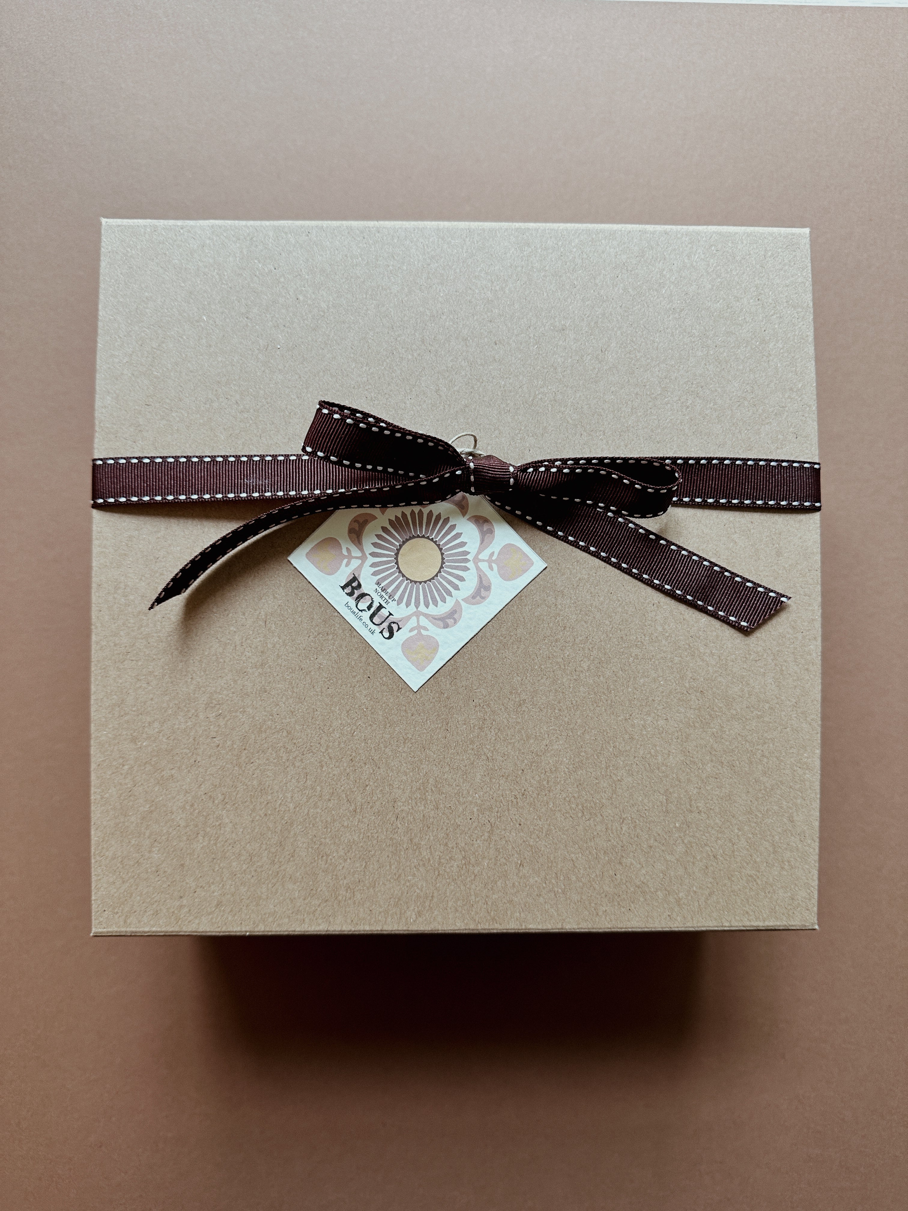Brown gift box with a brown ribbon and a small card on a beige background