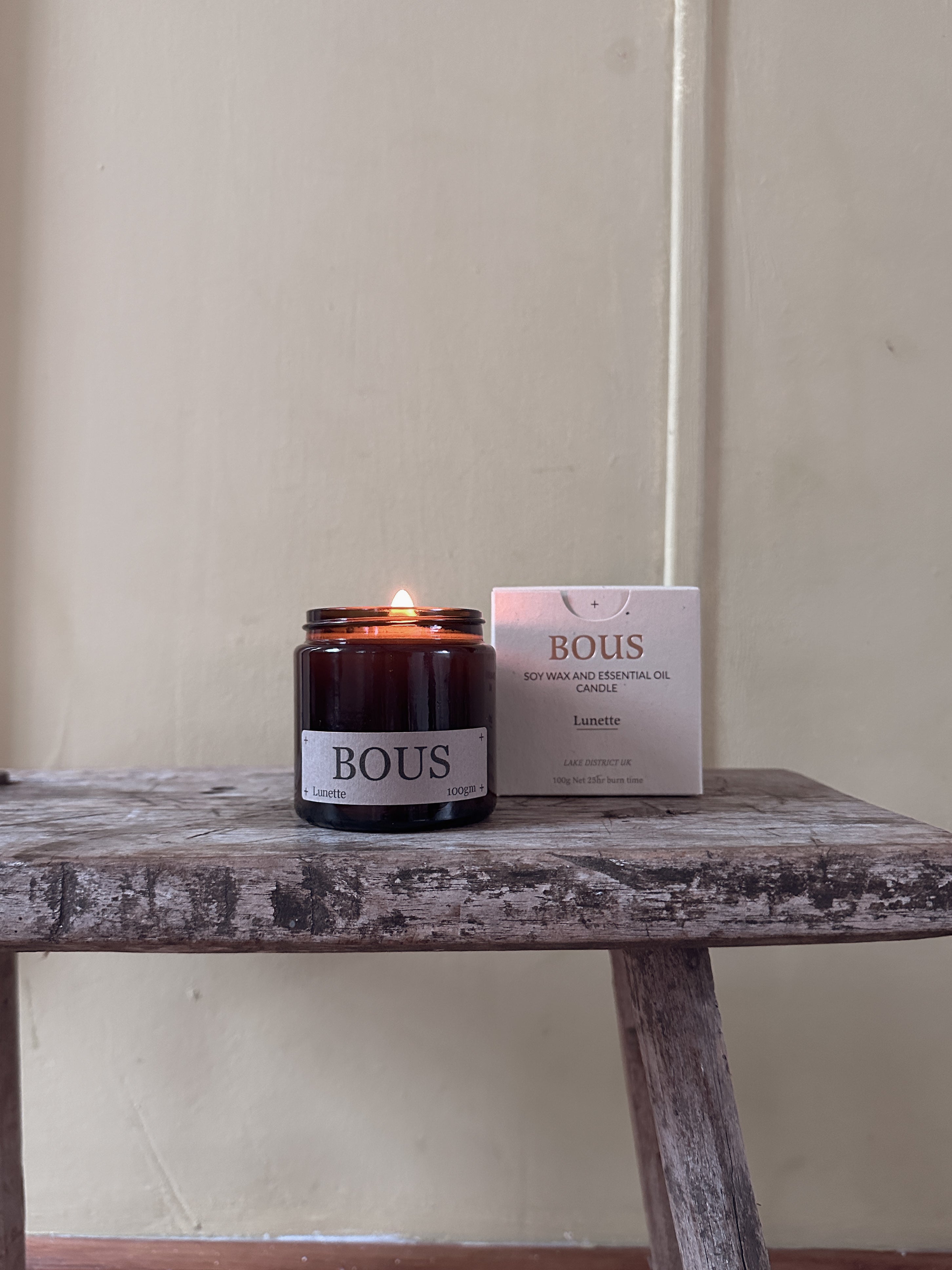 Candle and packaging labeled 'BOUS' on a wooden table against a plain wall.